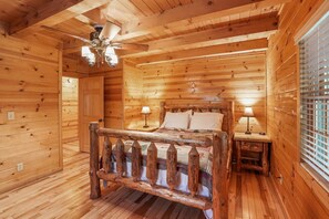 A wooden cabin bedroom with a bed, two bedside lamps, a ceiling fan, and a window with blinds.