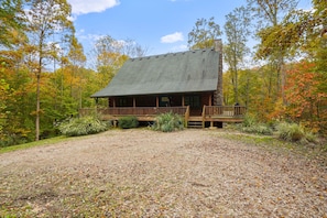 A wooden cabin with a porch, surrounded by a gravel driveway and autumn foliage. - Sieri Glen Cabin | Hocking Hills, Sleeps 14 (Logan)