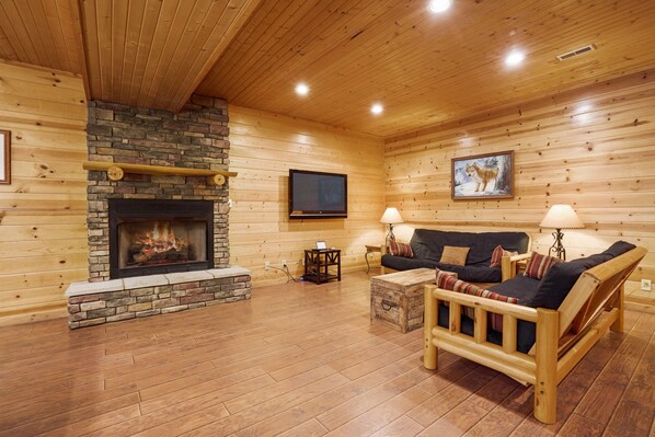 A log cabin interior with a stone fireplace, wooden furniture, and a television mounted on the wall.