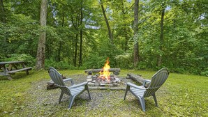 A campsite with a fire pit, two chairs, and a picnic table surrounded by a forest. - Sieri Glen Cabin | Hocking Hills, Sleeps 14 (Logan)