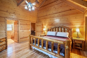A wooden cabin bedroom with a bed, two bedside tables, a ceiling fan, and a door leading to another room. - Sieri Glen Cabin | Hocking Hills, Sleeps 14 (Logan)