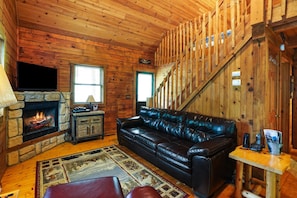 A cozy cabin interior with a stone fireplace, leather sofa, and a wooden staircase.