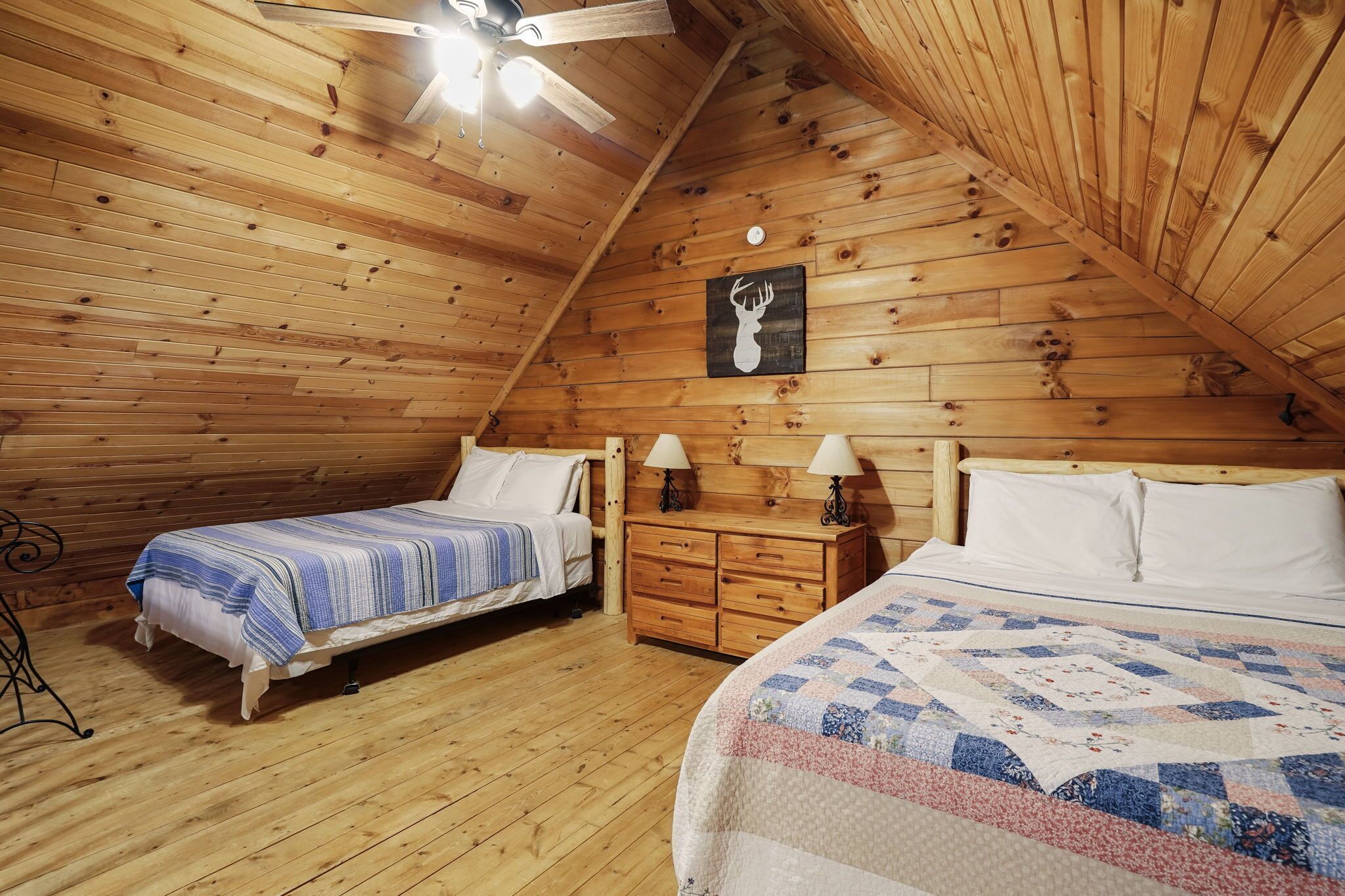 Un dormitorio estilo ático acogedor con dos camas, paredes de madera, ventilador de techo y una lámpara de pared.