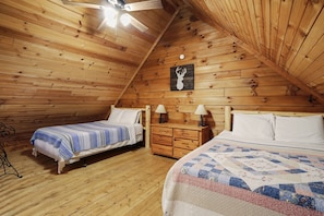 A cozy attic bedroom with wooden walls, a sloped ceiling, two beds, a dresser, and a deer head artwork.
