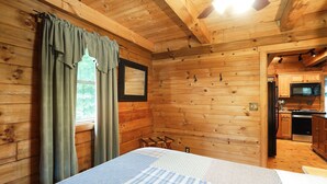 Un dormitorio de cabaña de madera con cama, una ventana con cortinas, una mesita y vistas a la cocina.