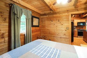 A wooden cabin bedroom with a bed, a window with curtains, a small table, and a view of the kitchen.