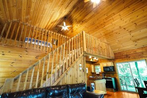 A wooden cabin interior with a staircase, a living area with a leather couch, and a kitchen area with a microwave.