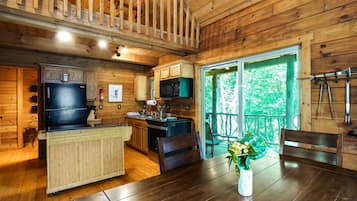 A wooden cabin interior with a kitchen, dining area, and a balcony with a view of trees.
