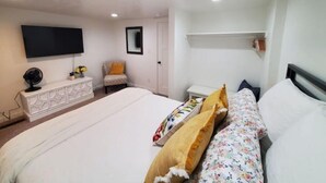 A bedroom with a bed, a TV mounted on the wall, a chair, a small table, and a shelf. - The White House Cottage (Inkom)