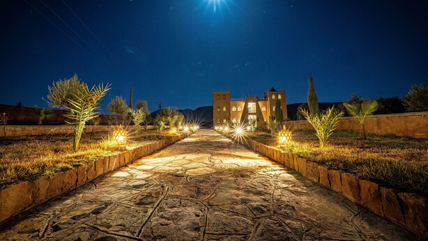 A stone pathway leading to a building with multiple windows, surrounded by grass and palm trees, under a night sky with a bright moon. - KASBAH NTIFA (Ait Taguella)