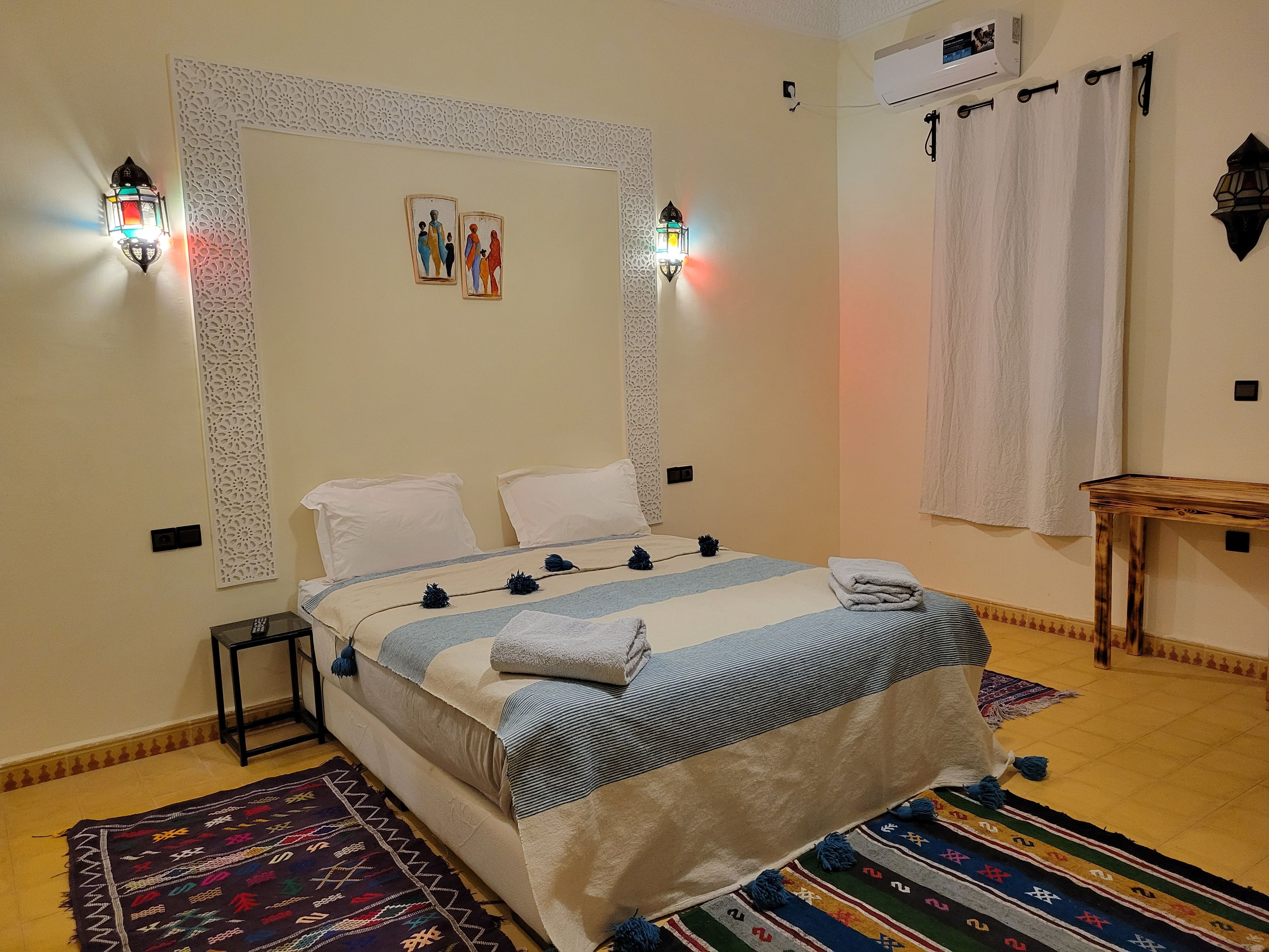 A hotel room with a bed, decorative wall panels, wall-mounted lights, a small table, and a window with curtains.