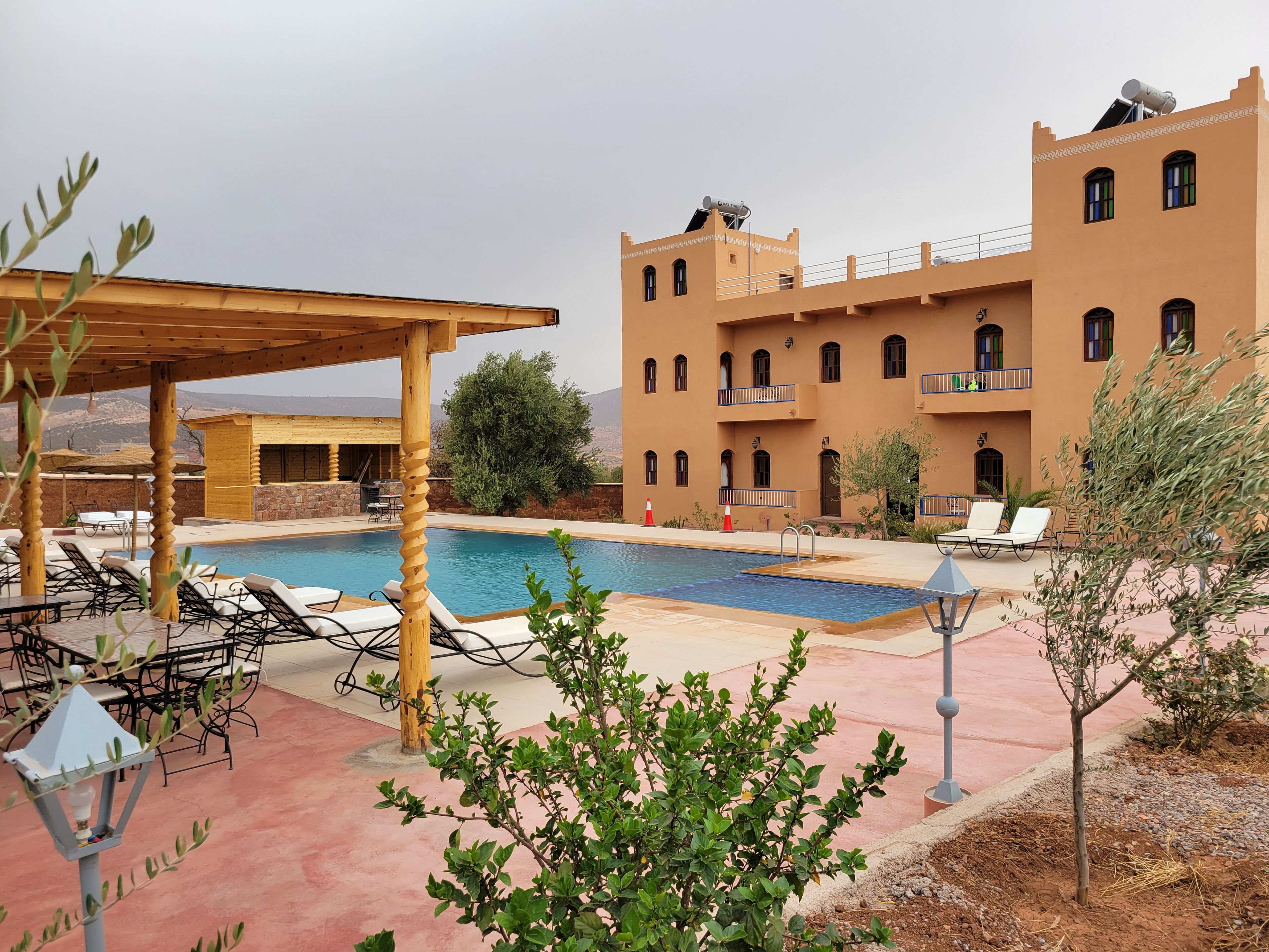A pool area with lounge chairs and a wooden pergola, a building with multiple balconies, and a clear view of the surrounding landscape.