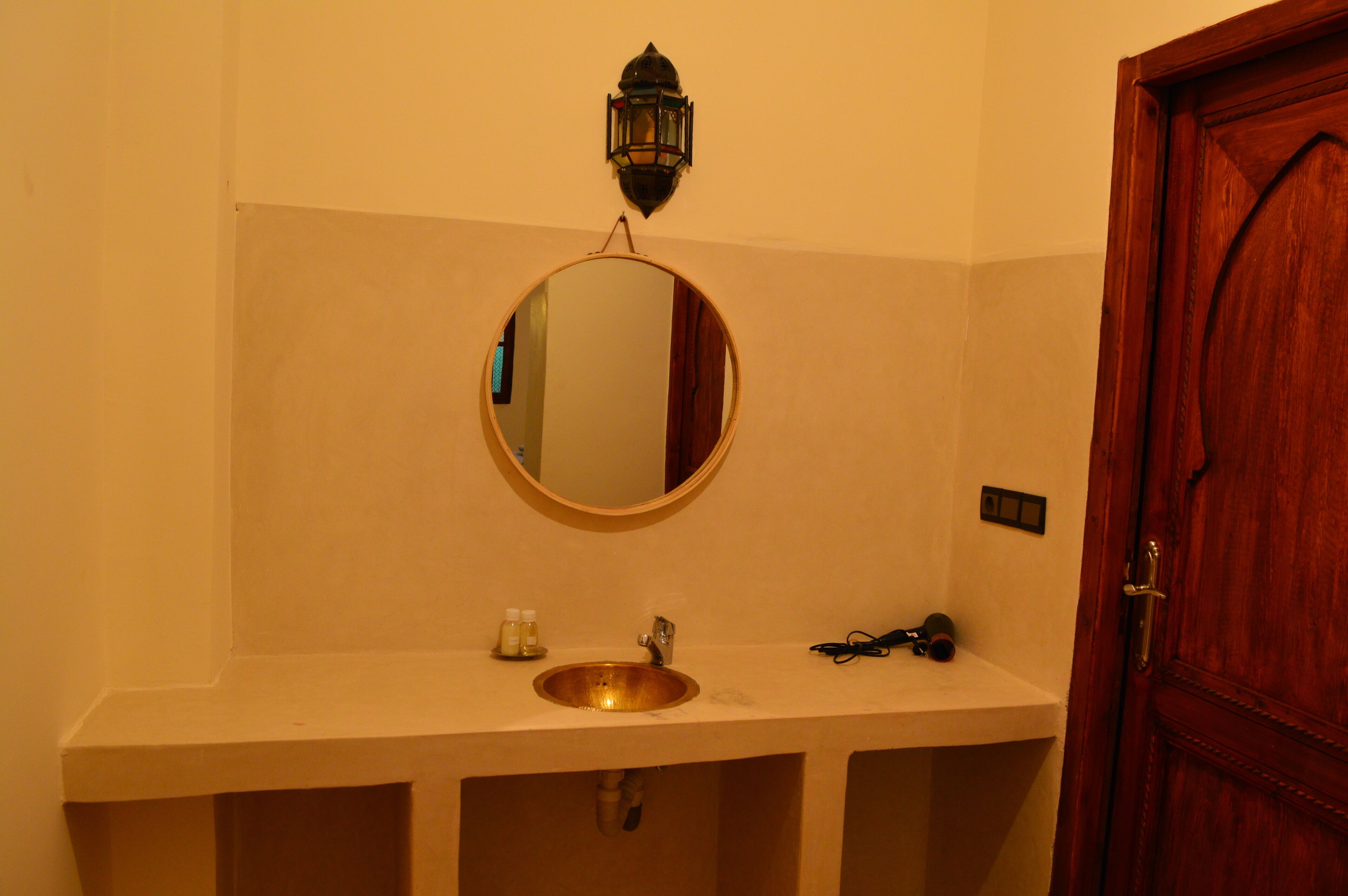 A bathroom with a round mirror, a sink, a small shelf with toiletries, and a wooden door.