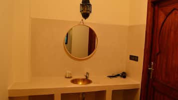 Un baño con un espejo redondo, un lavabo, un estante pequeño con artículos de aseo y una puerta de madera.