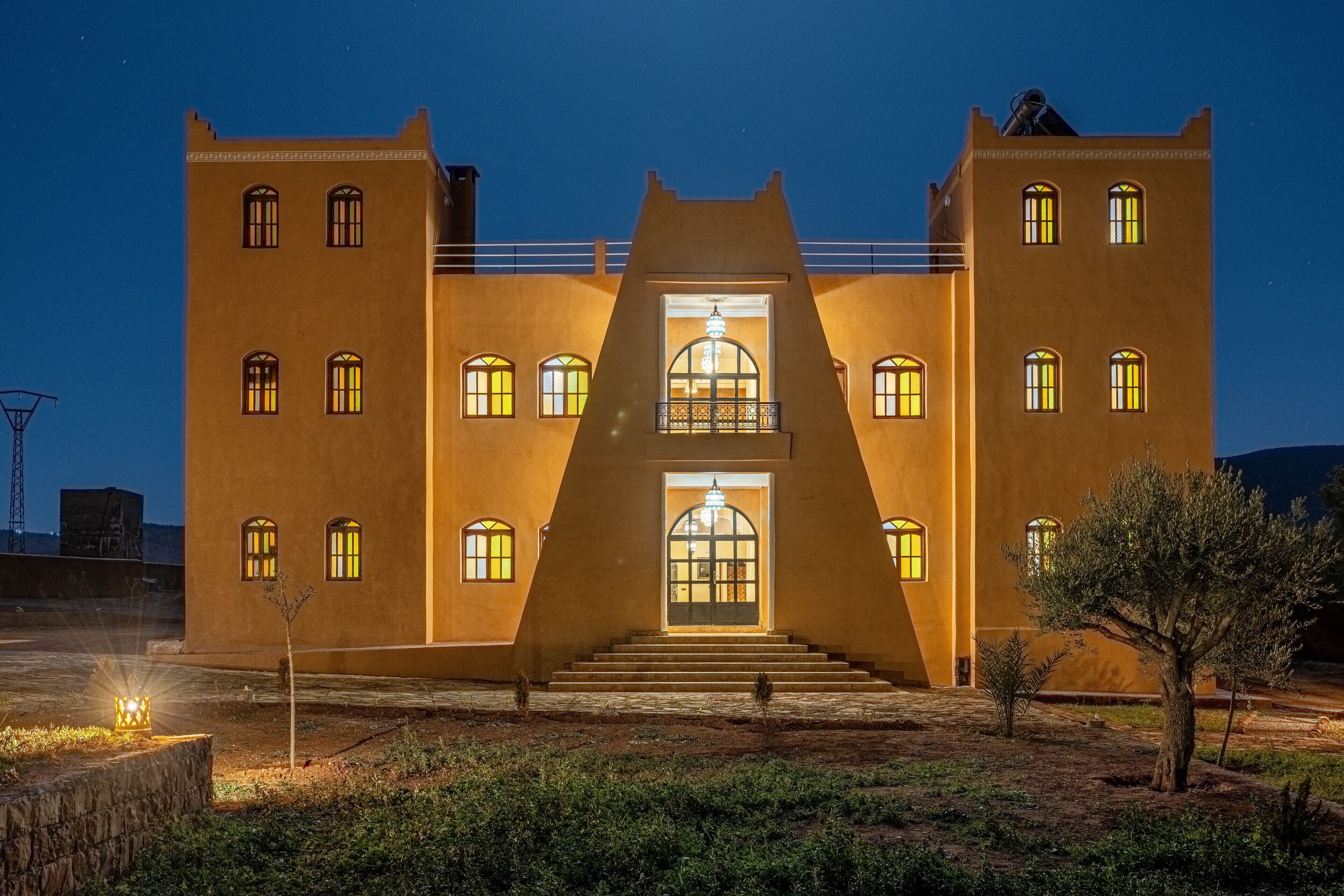 A large, symmetrical building with a central staircase leading to a balcony, surrounded by a landscaped area with sparse vegetation.