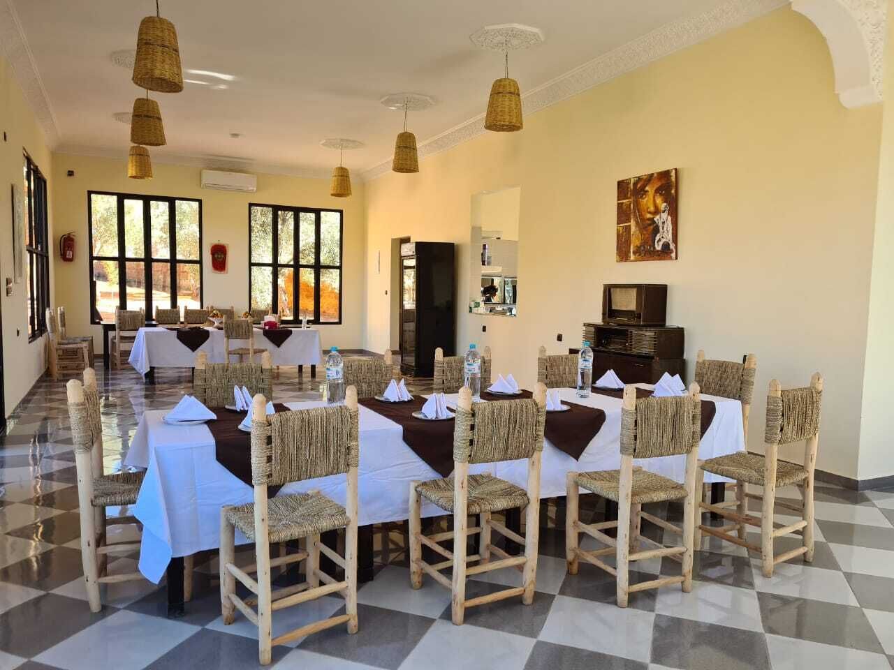 Un espace repas avec plusieurs tables et des chaises en osier, de grandes fenêtres et un sol à carreaux.