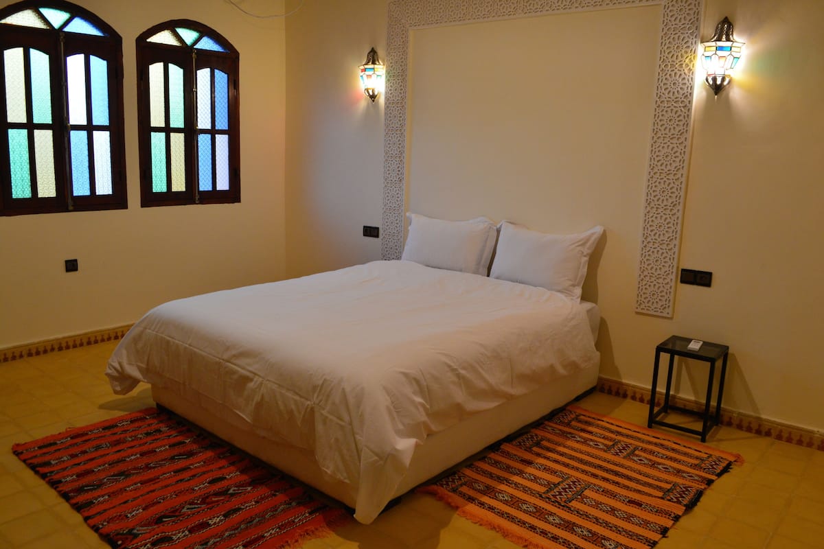 A neatly made bed with white linens, two wall-mounted light fixtures, and a patterned rug on the floor.