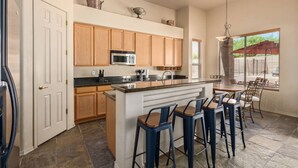 A kitchen with wooden cabinets, a central island with bar stools, a microwave, and a dining area with a table and chairs. - Troon/Gated/Pool/Firepit/Sleep 6 (Scottsdale)