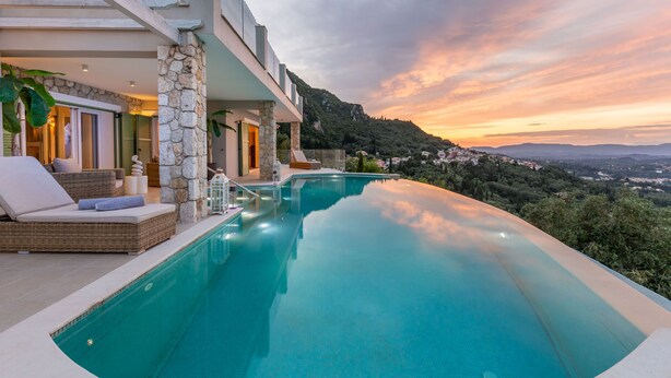 Una piscina infinita moderna con pared de piedra y mobiliario de mimbre, con vistas a un paisaje montañoso al atardecer.
