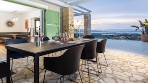 Un área de comedor al aire libre con suelo de piedra, una mesa dispuesta con platos, vasos y una botella, y vistas a la ciudad y las montañas al fondo.