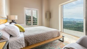 Un dormitorio con cama, mesita de noche, lámpara y vista a un paisaje por una ventana grande.