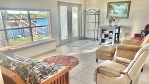 A living room with a view of a dock and water, a wooden bench, beige armchairs, and a small table with a vase. - Salty River Lodge Arroyo City, TX (Cameron County)