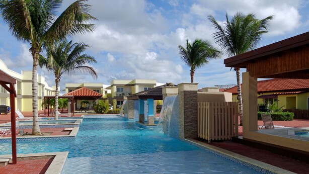 A swimming pool with a waterfall feature, surrounded by palm trees and lounge chairs.