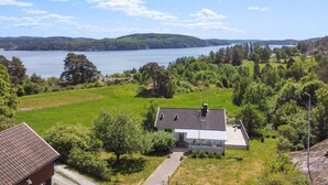 Una casa con techo inclinado y un pequeño porche, rodeada de un exuberante paisaje verde y un lago.