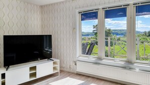 Una habitación con televisor sobre un soporte blanco, un radiador y un ventanal con vistas a zonas verdes y colinas.