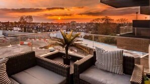 Eine Dachterrasse mit Korbmöbeln, einem Blumentopf und Blick auf einen Sonnenuntergang über der Stadt.