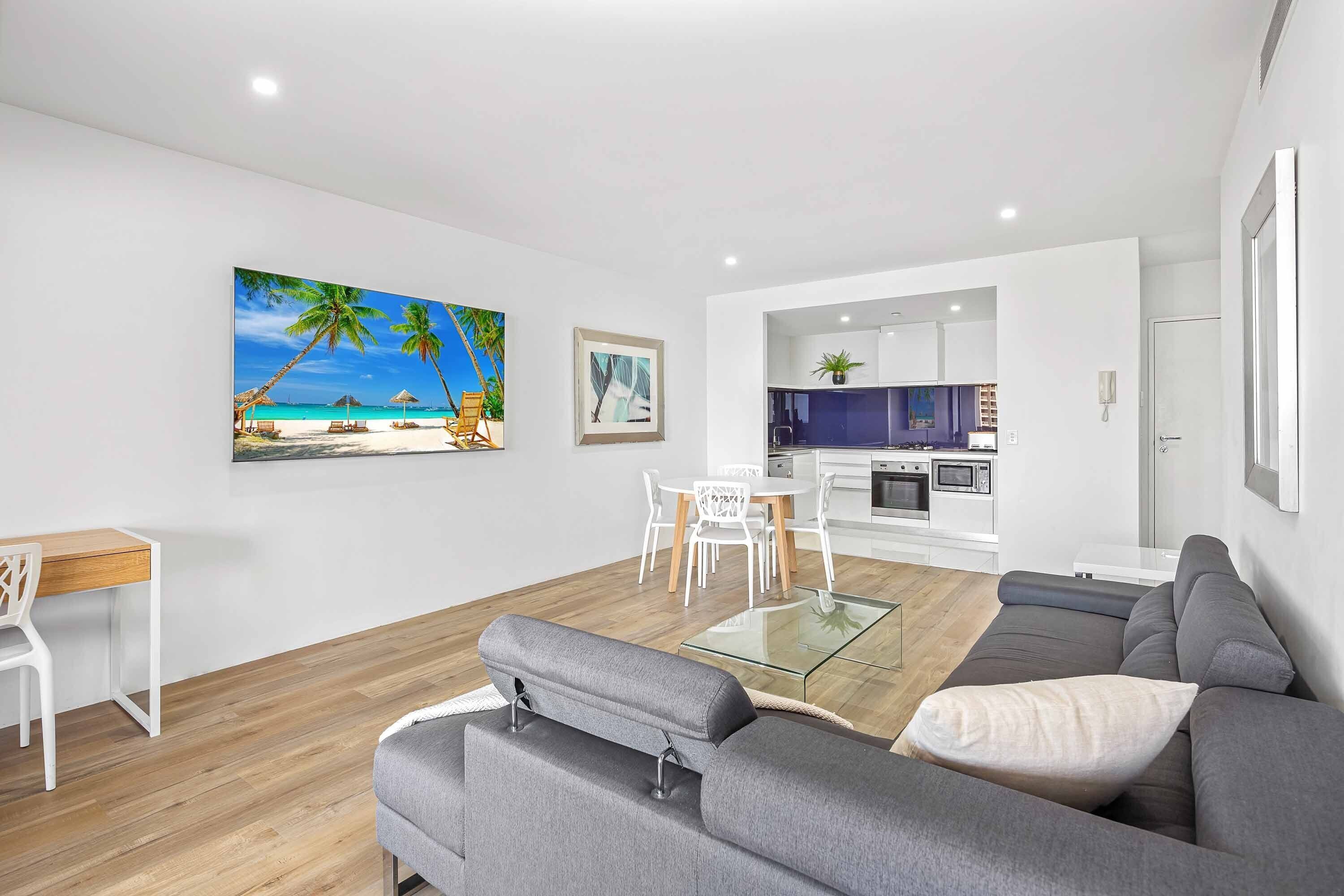 Een moderne woonkamer met een grijze bank, een glazen salontafel en een eetgedeelte met witte stoelen. In de achtergrond is een keuken zichtbaar met witte kasten en een blauw aanrecht.