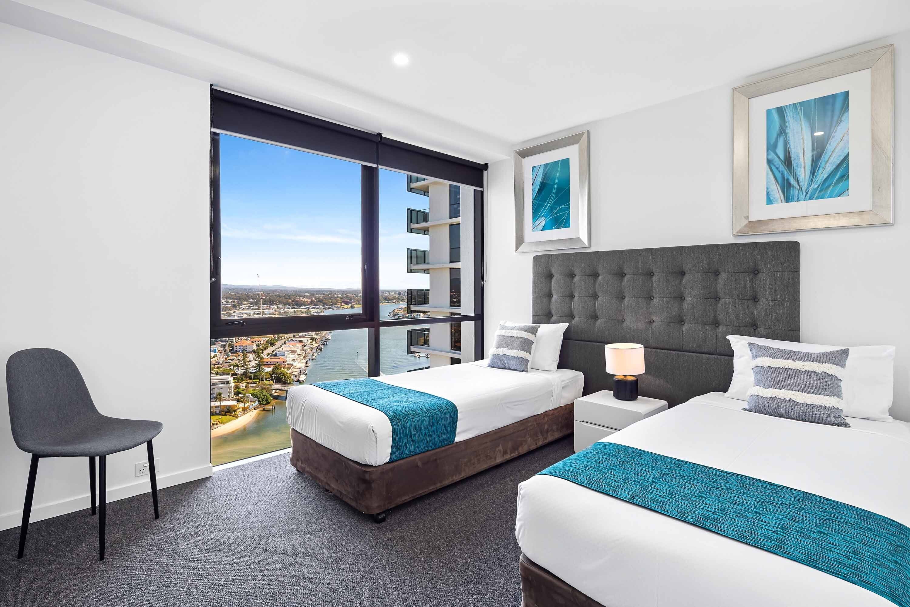 A modern hotel room with two beds, a large window with a city view, and a chair.
