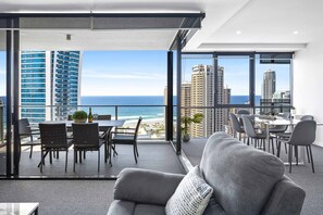 Ein modernes Wohnzimmer mit grauem Sofa, einer Essecke mit Tisch und Stühlen sowie einem Balkon mit Blick auf das Meer und die Stadtlandschaft.