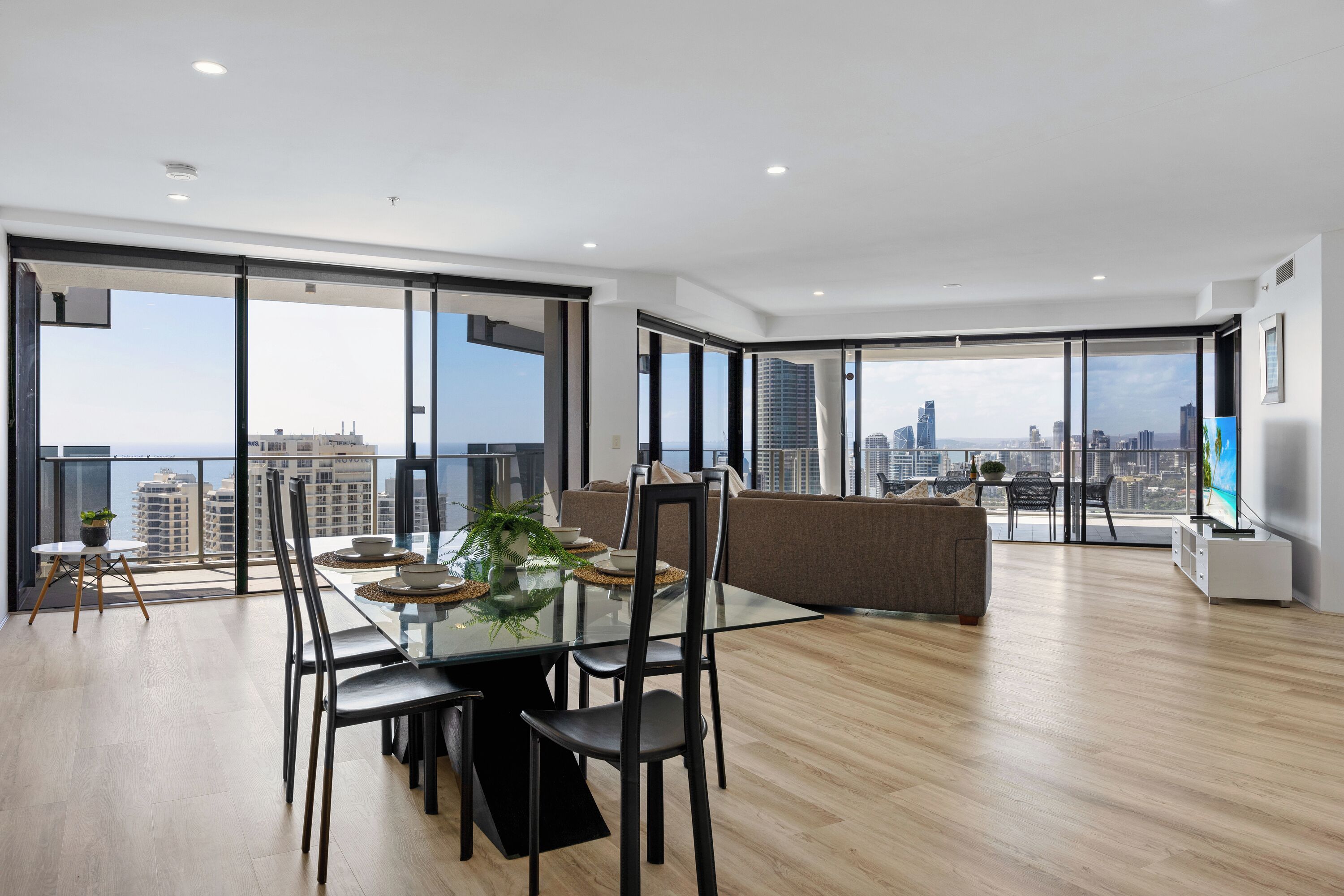 A spacious living area with a dining table, chairs, and a sofa. There is a large window offering a city view.