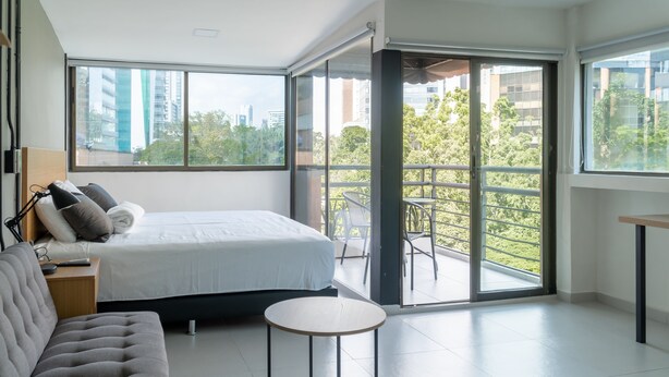 A modern bedroom with a large bed, a sofa, a small round table, and a balcony with a view of the city. - HOTEL BAMBU MILLA DE ORO (Medellín)