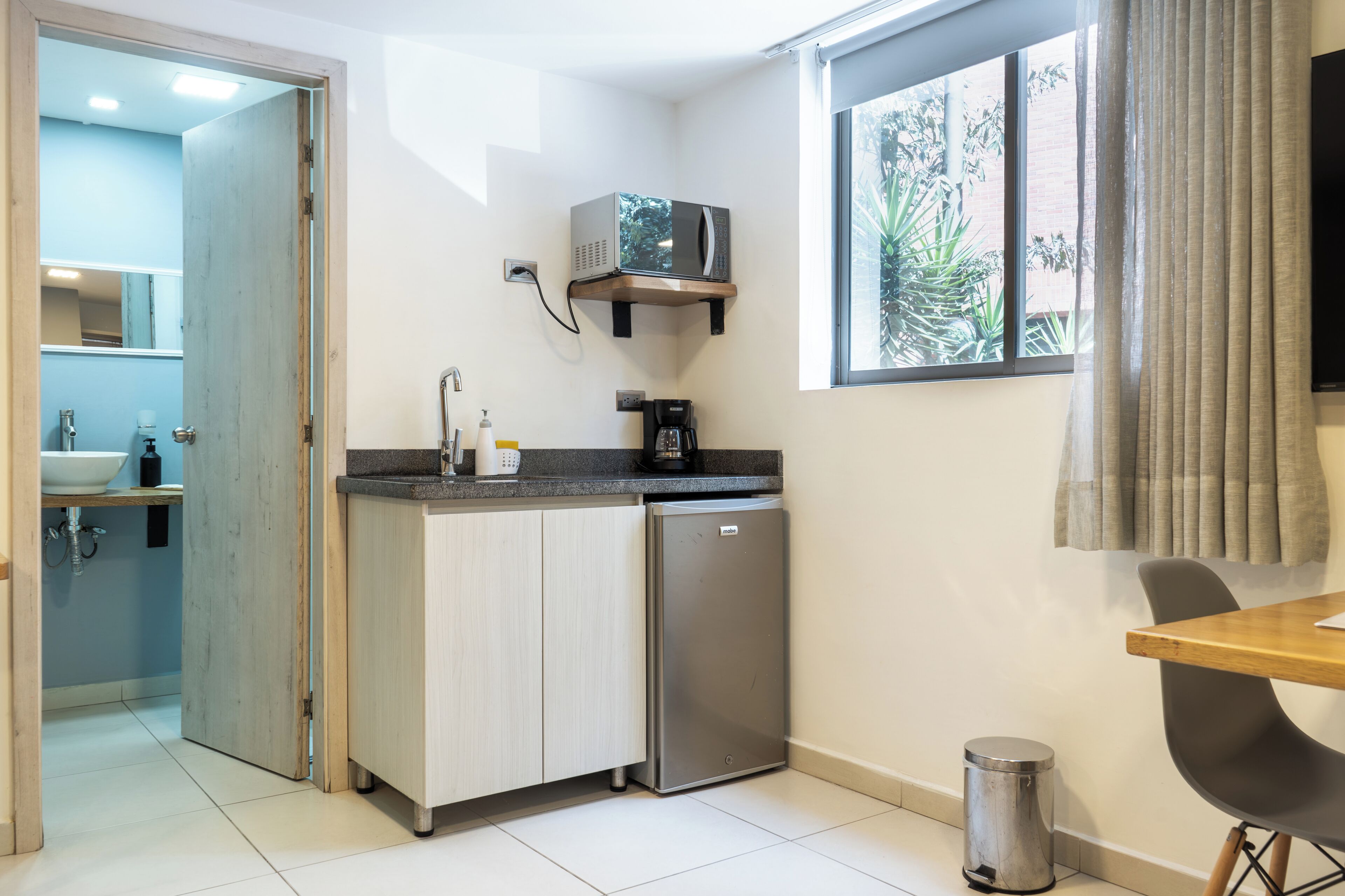 Eine kompakte Küche mit Mikrowelle, Kaffeemaschine und kleinem Kühlschrank. Ein Fenster sorgt für natürliches Licht, und ein Mülleimer ist sichtbar.