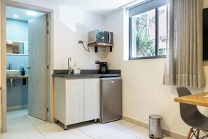 Eine kompakte Küche mit Mikrowelle, Kaffeemaschine und kleinem Kühlschrank. Ein Fenster sorgt für natürliches Licht, und ein Mülleimer ist sichtbar.