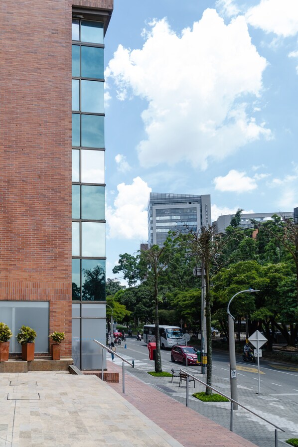 A modern building with a brick facade and large windows, a paved sidewalk, and a street with vehicles and trees in the background.