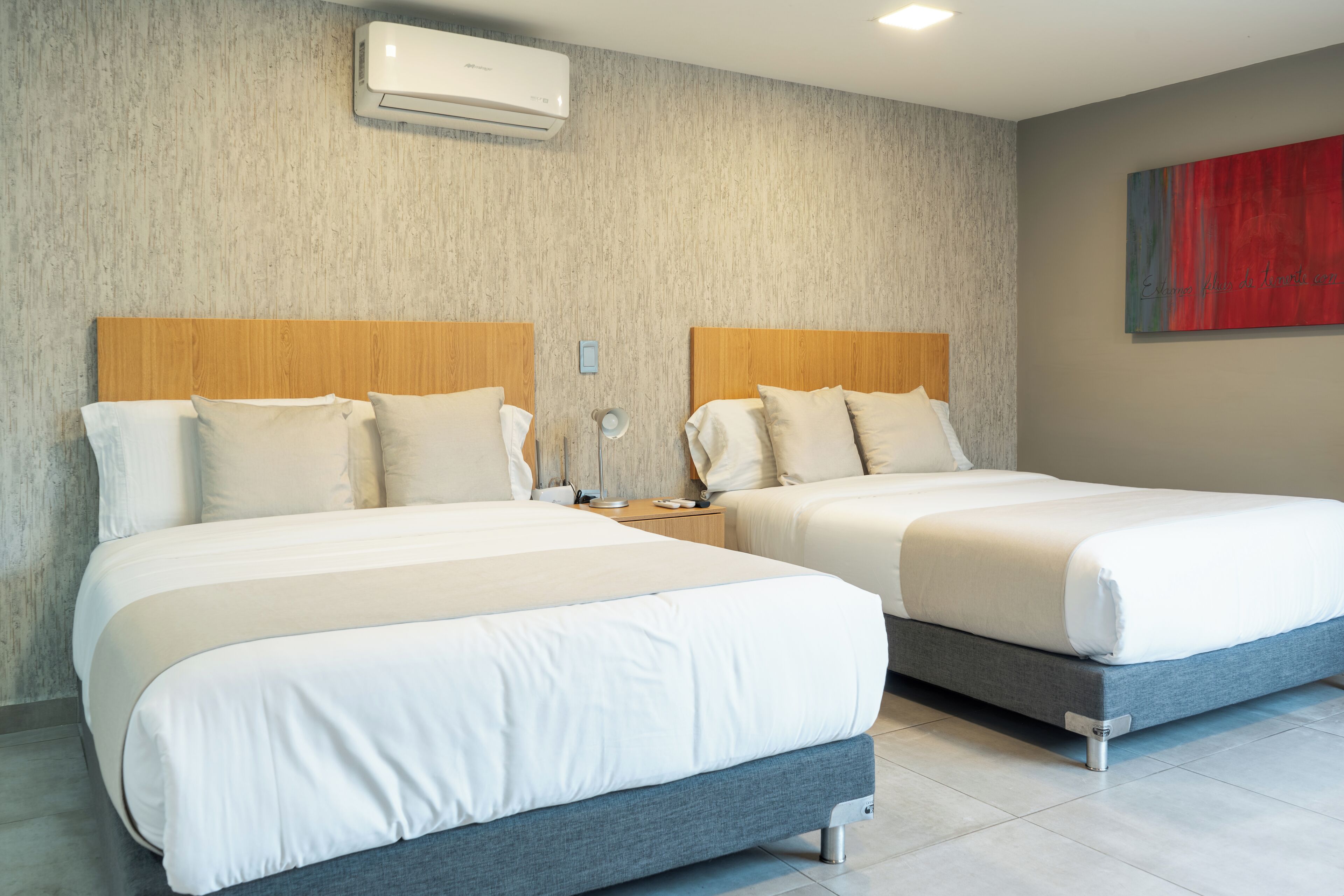 Une chambre d’hôtel avec deux lits, une tête de lit en bois, un climatiseur fixé au mur et un tableau abstrait rouge.