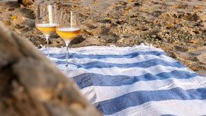 A beach scene with two glasses of a beverage on a striped towel, a large rock in the foreground, and a calm sea in the background. - Casa Eos - prime location property (Santa Teresa de Cobano)