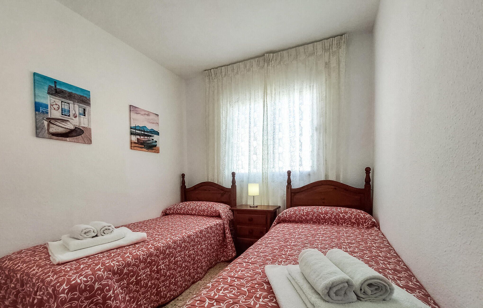 Una habitación pequeña y acogedora con dos camas individuales, una mesita de noche y una ventana con cortinas traslúcidas.