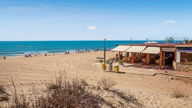 Un restaurante frente a la playa con terraza, vista al océano y una torre de salvavidas al fondo.