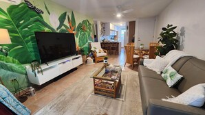 Ein Wohnzimmer mit großem Flachbildfernseher, einer Couch, einem Couchtisch und einem Essbereich im Hintergrund.