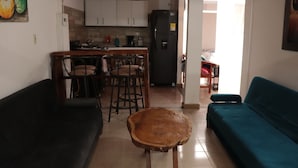 Un salon avec un canapé noir, un canapé de couleur turquoise et une table basse en bois. On y trouve également un coin cuisine avec des meubles blancs et un réfrigérateur noir.