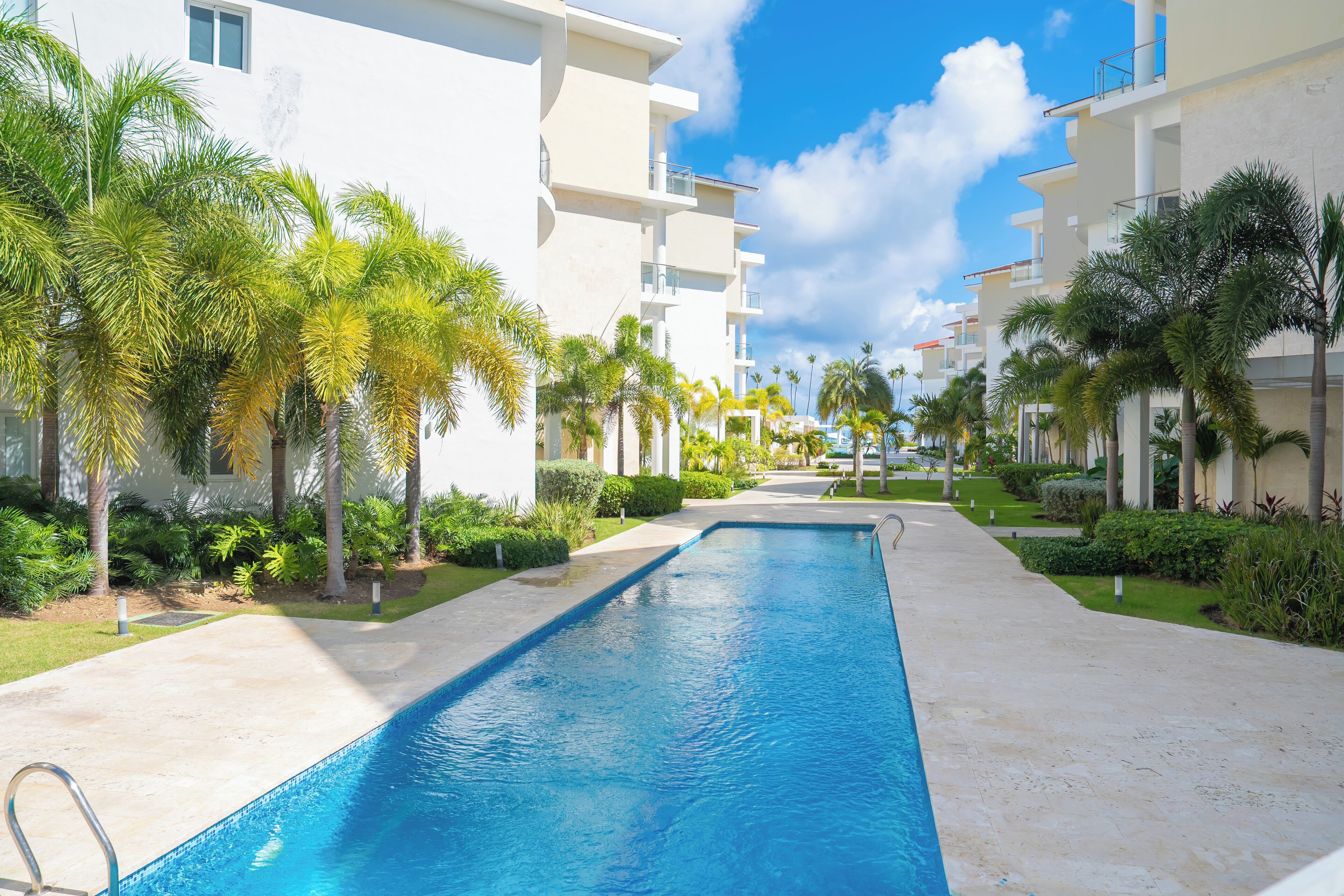 A swimming pool with a clear blue water surface, surrounded by a pathway lined with palm trees and greenery, leading to a multi-story building with balconies.