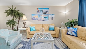 Ein Wohnzimmer mit einem beigen Sofa, einem blauen Sessel und einem Couchtisch. Zwei Beistelltische mit Lampen und Dekorationselementen stehen ebenfalls im Raum. Eine Deckenleuchte und eine Wand mit drei gerahmten Bildern runden das Bild ab.