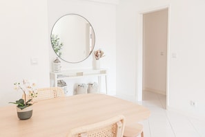 A dining area with a round mirror, white walls, a wooden table, and wicker chairs.