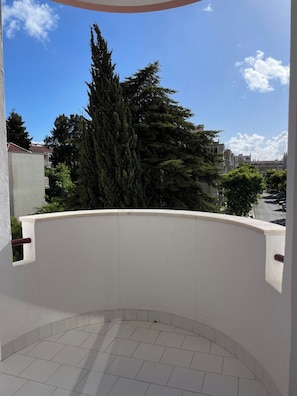 A balcony with a tiled floor, overlooking a street with trees and buildings.