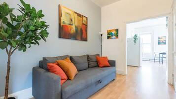 A modern living room with a grey sofa, orange cushions, and a potted plant. There are abstract paintings on the walls and a doorway leading to another room.