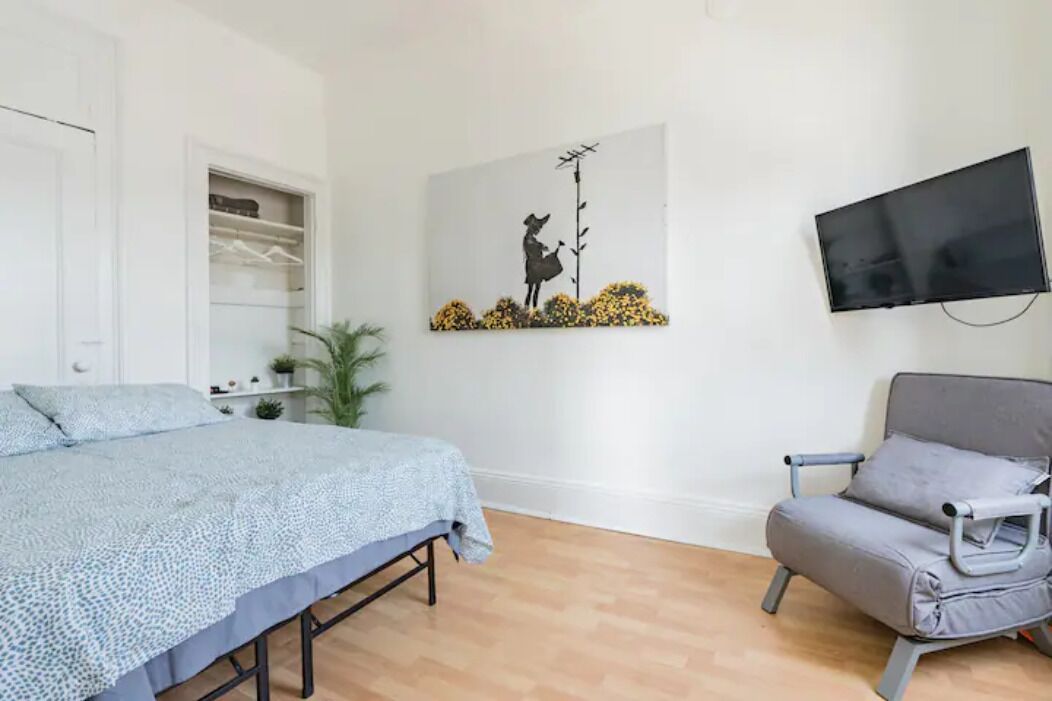 Um quarto com cama, um sofá cinza, uma TV fixada na parede e um quadro de uma pessoa com um pássaro.